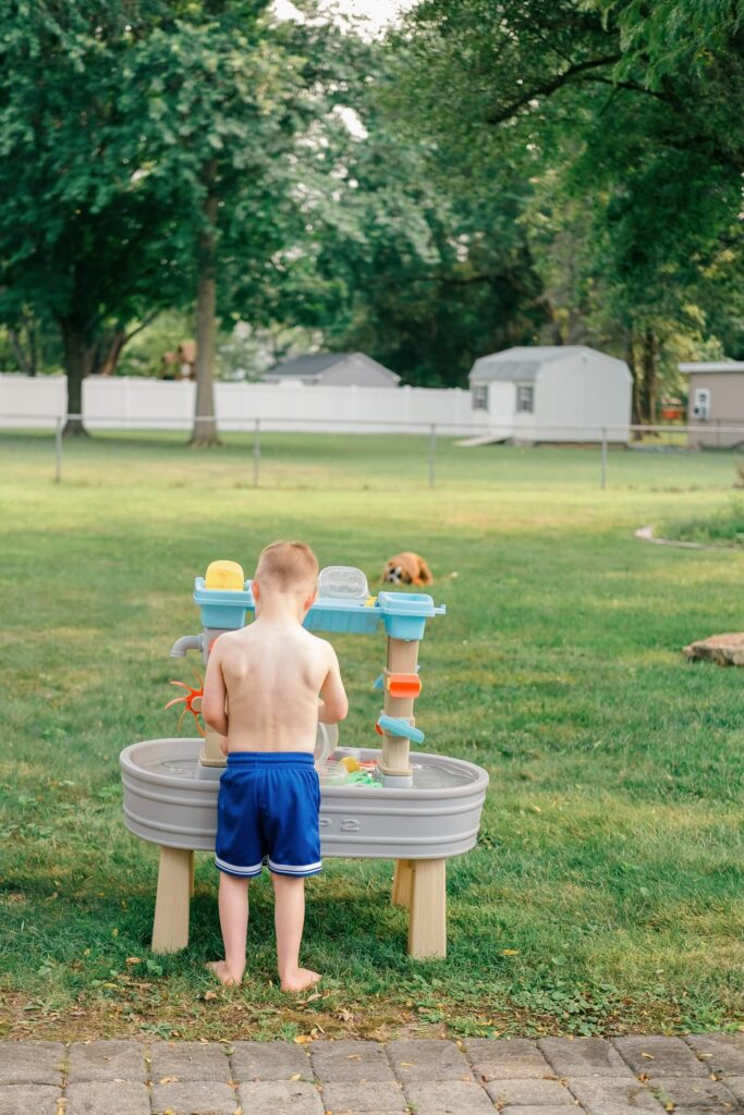 Your August Photo Scavenger Hunt A young boy playing at an outdoor water table in the backyard, captured as part of a summer photo scavenger hunt